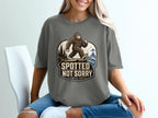 Bigfoot with sunglasses walks on a cliff above waves; shirt text: Spotted Not Sorry.
