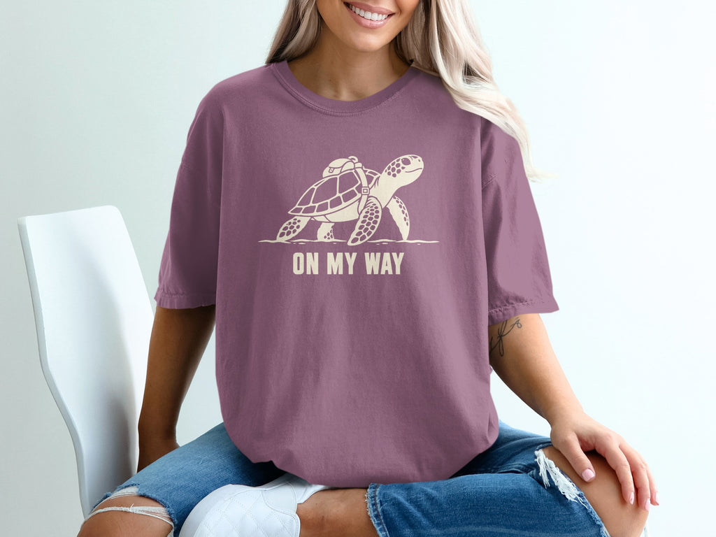 Purple t-shirt with a cream graphic of a turtle wearing a backpack and text "ON MY WAY".