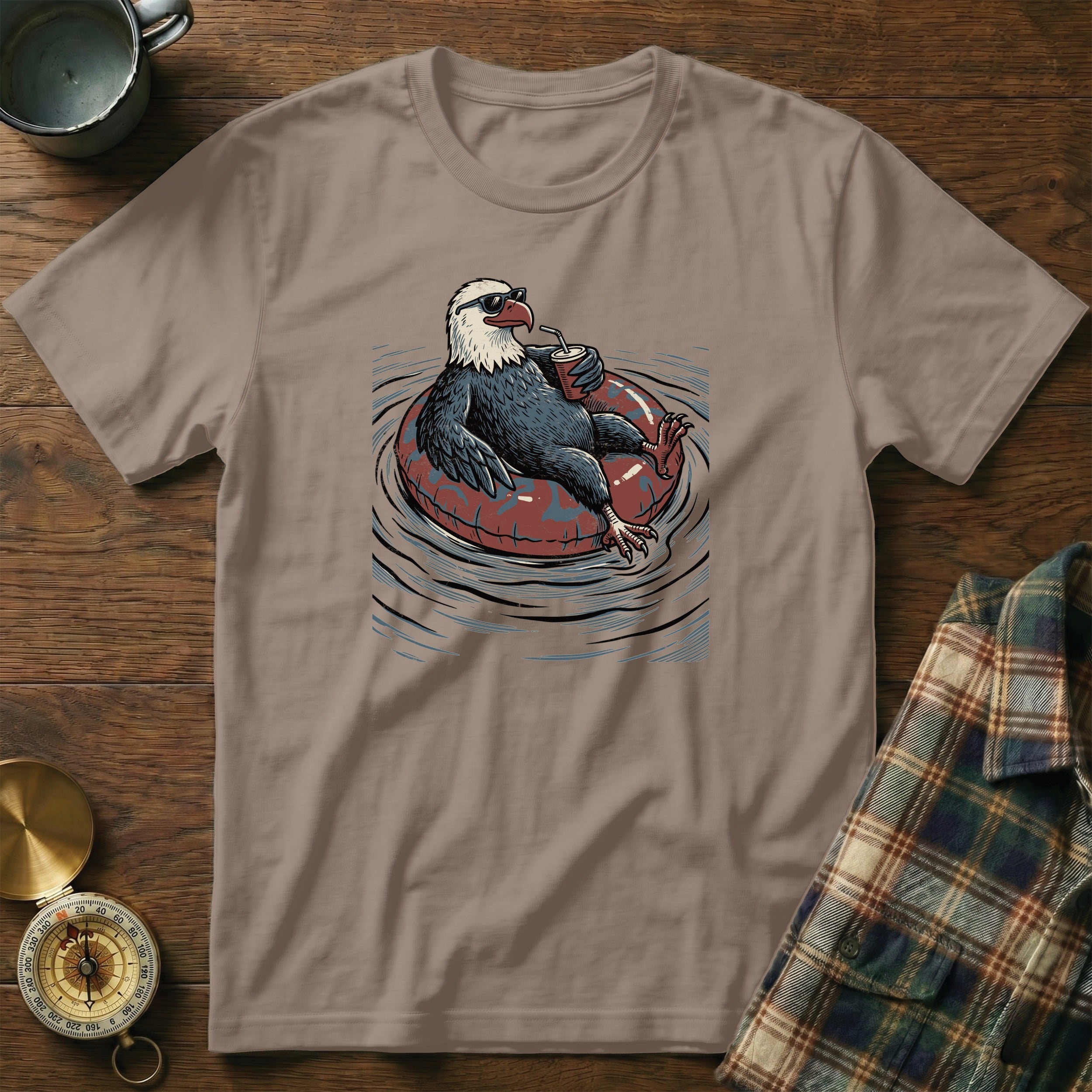 Graphic t-shirt with an eagle wearing sunglasses floating on an inner tube drinking a beverage.