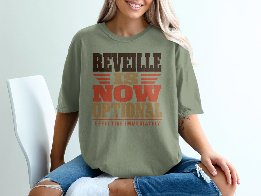 A woman wears an olive green t-shirt featuring the distressed brown, tan, and red text "REVEILLE IS NOW OPTIONAL EFFECTIVE IMMEDIATELY".