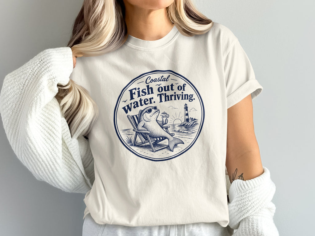 A person wearing an off-white t-shirt featuring a circular graphic of a fish lounging in a beach chair with the text Coastal Fish out of water. Thriving.