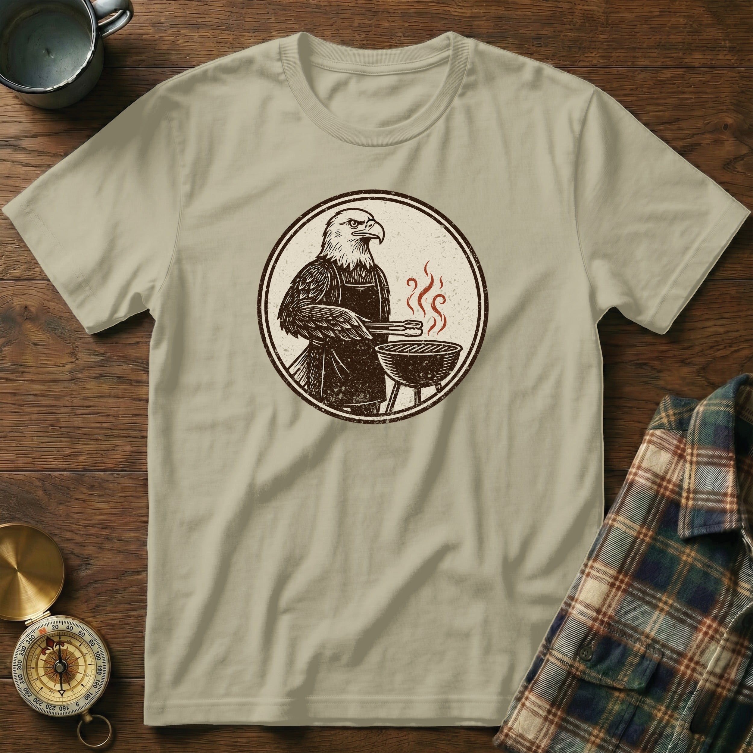Graphic of eagle in apron grilling, on a tan t-shirt.
