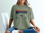 Olive green t-shirt graphic with a canoe on a lake at sunset.