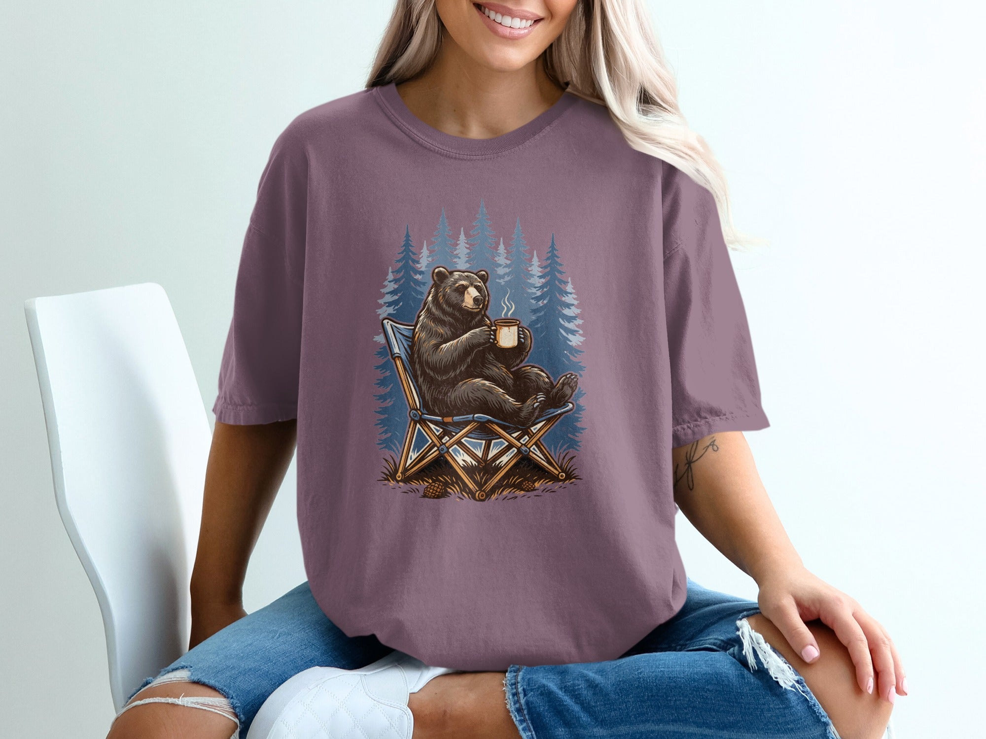 Bear sitting in chair holding coffee mug with pine trees background.