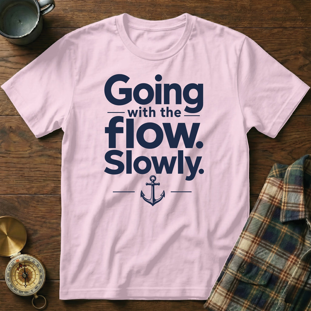 A light pink t-shirt with navy blue text reading Going with the flow. Slowly., centered above an anchor graphic, resting on a wooden surface.