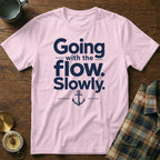 A light pink t-shirt with navy blue text reading Going with the flow. Slowly., centered above an anchor graphic, resting on a wooden surface.