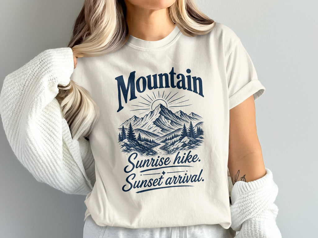 A person wearing an off-white t-shirt printed with a blue mountain graphic and the text Mountain Sunrise hike, Sunset arrival.