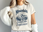 A person wearing an off-white t-shirt printed with a blue mountain graphic and the text Mountain Sunrise hike, Sunset arrival.