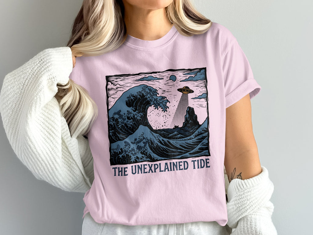 Pink t-shirt with a graphic of a UFO over crashing waves and the text "THE UNEXPLAINED TIDE".