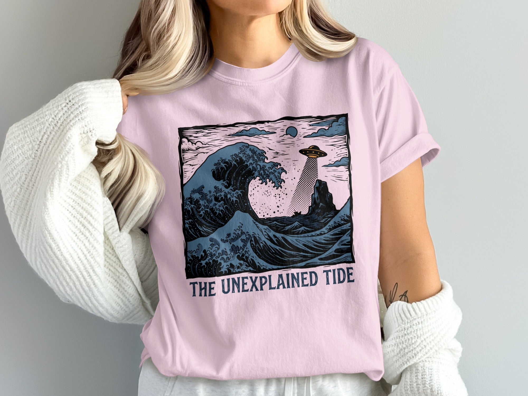 Pink t-shirt with a graphic of a UFO over crashing waves and the text "THE UNEXPLAINED TIDE".