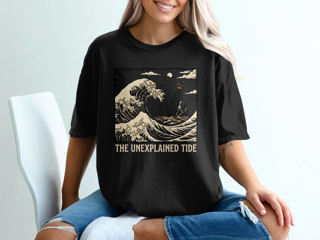 Black t-shirt with graphic of a large wave, small boats, and text "THE UNEXPLAINED TIDE".
