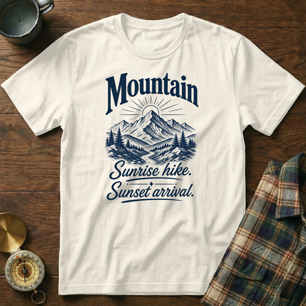 An off-white short-sleeve t-shirt features a blue graphic of mountains and the text "Mountain Sunrise hike. Sunset arrival." on a wooden background.