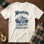 An off-white short-sleeve t-shirt features a blue graphic of mountains and the text "Mountain Sunrise hike. Sunset arrival." on a wooden background.