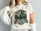 White t-shirt with shield graphic featuring mountains, trees, lake, and text: "NOWHERE NEAR DONE EXPLORING."