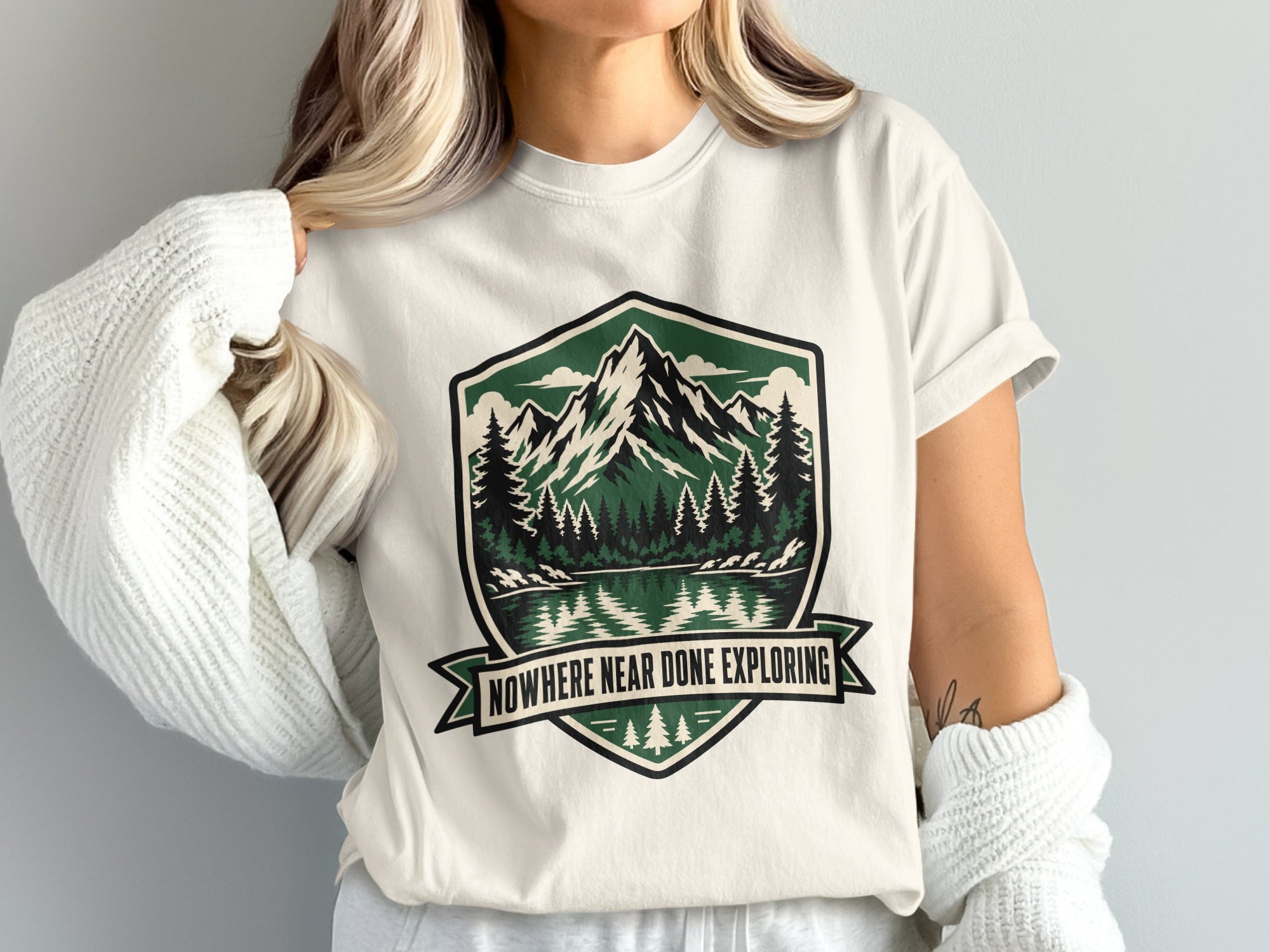 White t-shirt with shield graphic featuring mountains, trees, lake, and text: "NOWHERE NEAR DONE EXPLORING."
