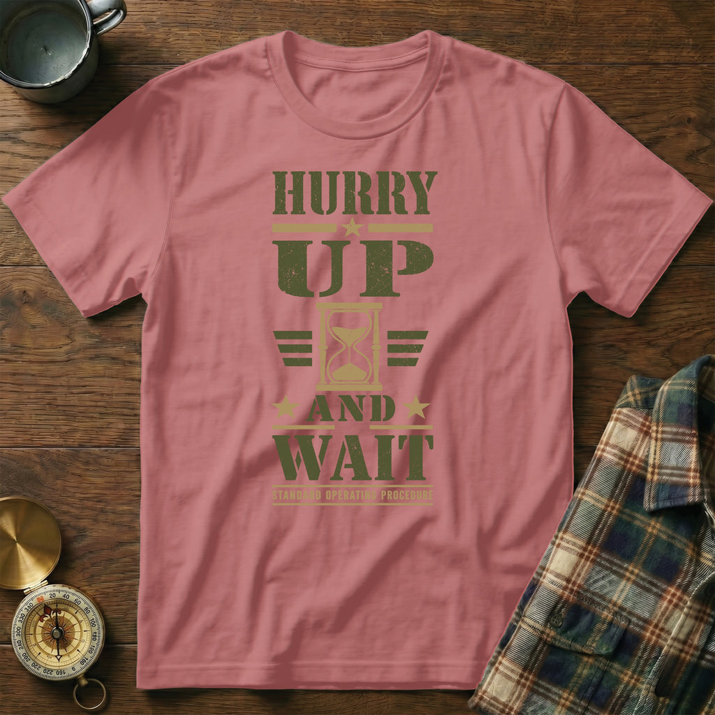 Dusty pink t-shirt featuring the text HURRY UP AND WAIT with an hourglass graphic, laid on a wooden surface.