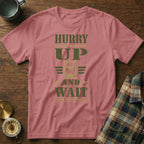 Dusty pink t-shirt featuring the text HURRY UP AND WAIT with an hourglass graphic, laid on a wooden surface.