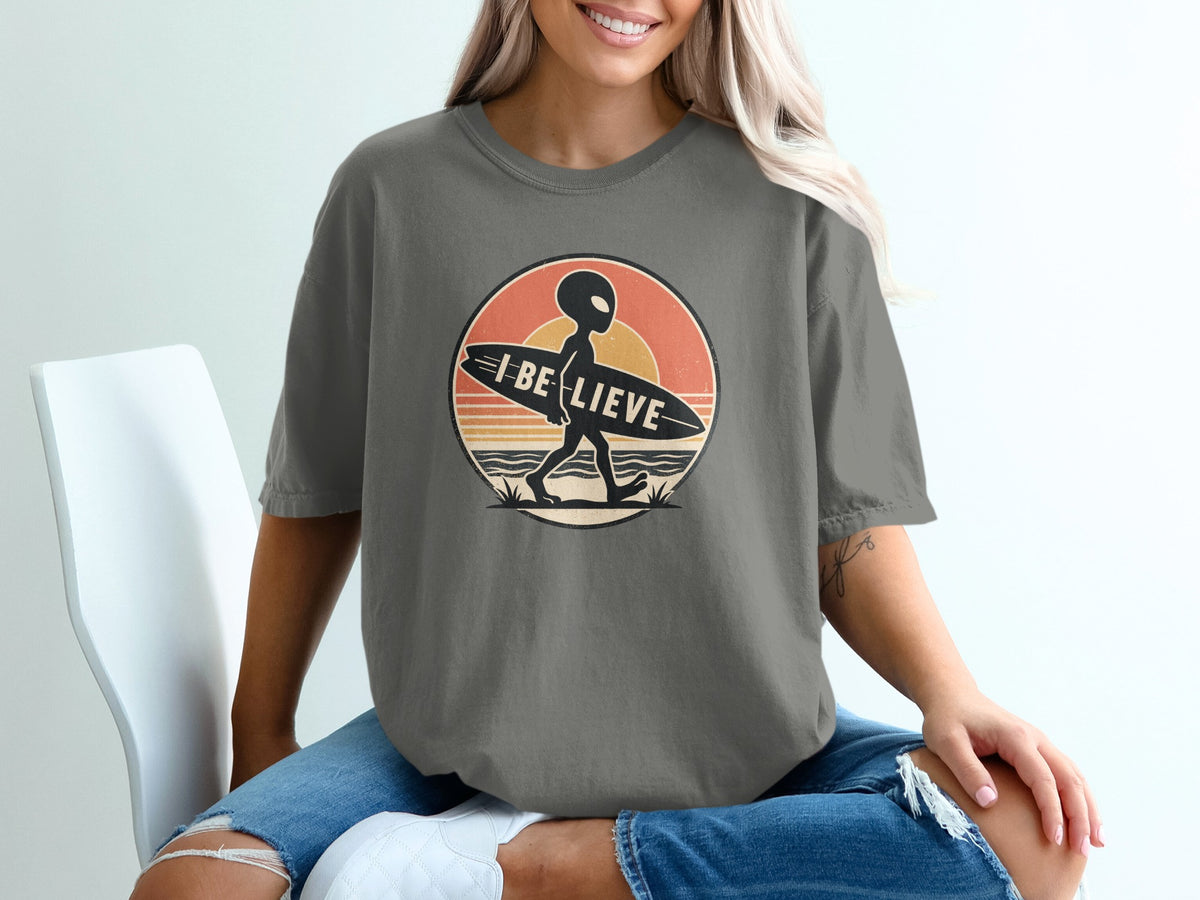 Alien carrying surfboard with "I Believe" text, against a retro sunset design.