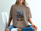 Bear sitting in a camping chair holding a hot drink, forest background graphic on shirt.