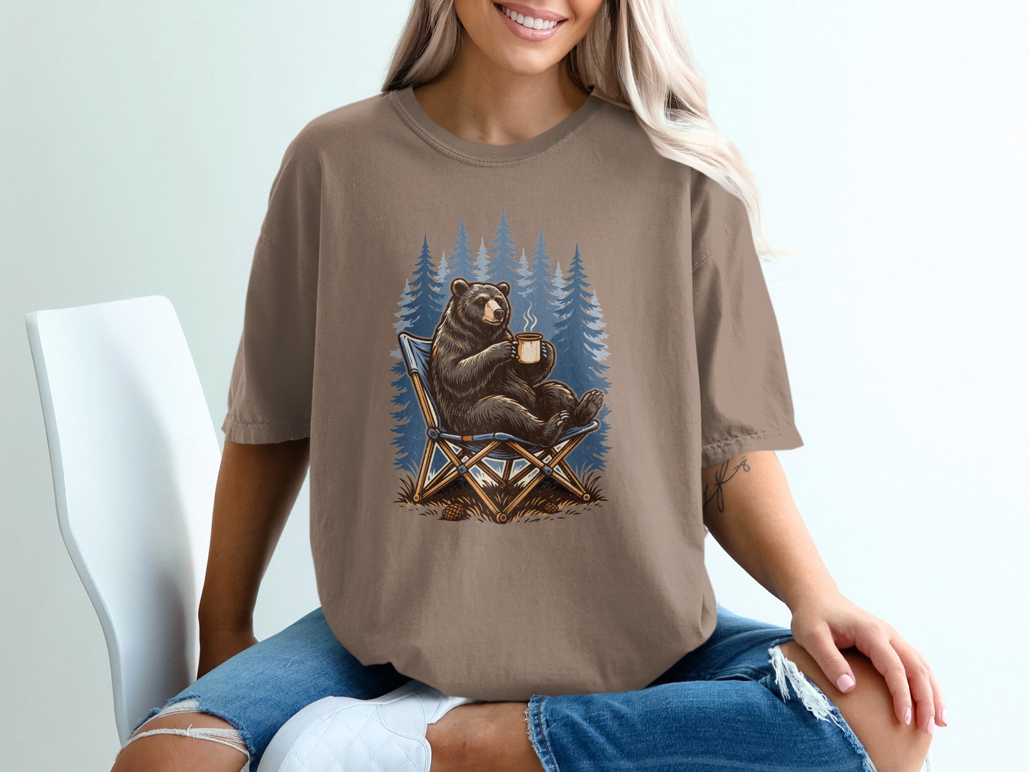 Bear sitting in a camping chair holding a hot drink, forest background graphic on shirt.