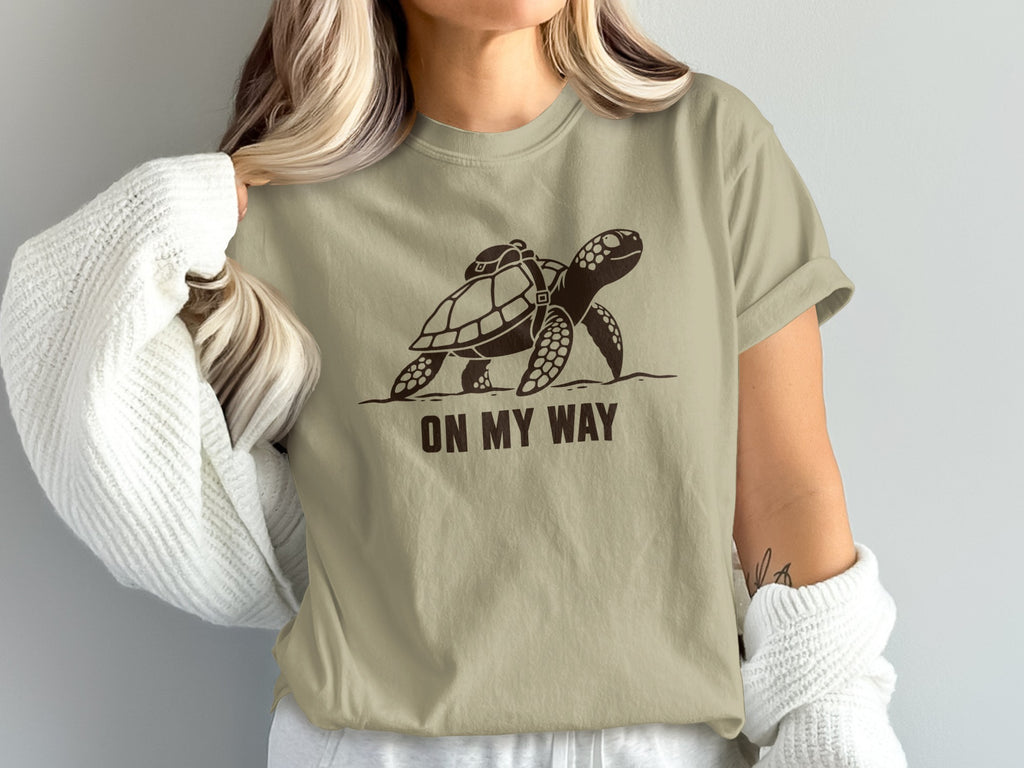 Brown graphic of a sea turtle with a backpack on a tan t-shirt, text reads "ON MY WAY".