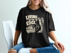 Black t-shirt with "Living on the Edge, Barely" text and a goat hanging from a cliff.