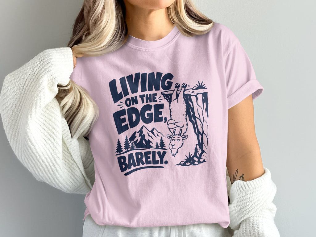 Pink t-shirt with navy graphic: "LIVING ON THE EDGE. BARELY." and a goat hanging from a cliff.