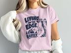 Pink t-shirt with navy graphic: "LIVING ON THE EDGE. BARELY." and a goat hanging from a cliff.