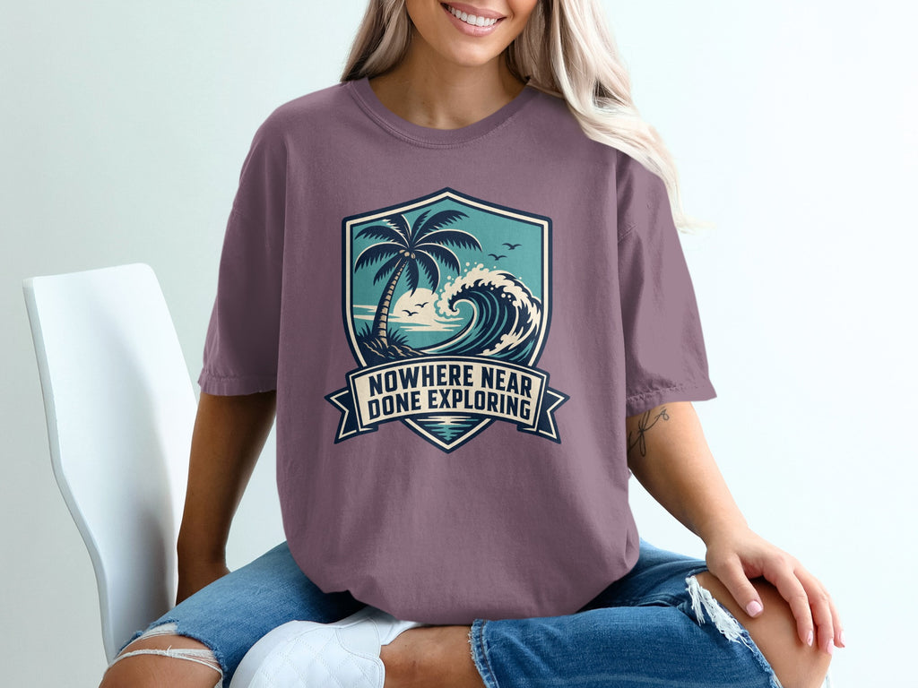 T-shirt with a crest graphic of a palm tree, ocean wave, and birds, text "Nowhere Near Done Exploring".