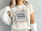 White t-shirt with "This Isn't Paradise You Just Visited We Stayed" text.