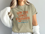 Beige t-shirt with text "LOW TIDE LOWER STANDARDS STILL SHOWING UP" and a crab graphic.