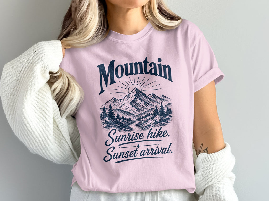 A blonde woman wears a pink crewneck t-shirt featuring a mountain graphic and the text Mountain Sunrise hike, Sunset arrival.