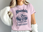 A blonde woman wears a pink crewneck t-shirt featuring a mountain graphic and the text Mountain Sunrise hike, Sunset arrival.