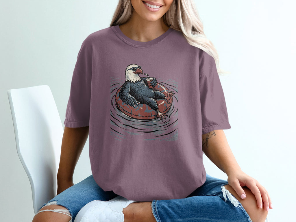 Eagle with sunglasses floats on inner tube with drink, printed on purple t-shirt.