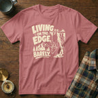 T-shirt with a goat hanging off a cliff, text "LIVING ON THE EDGE, BARELY."