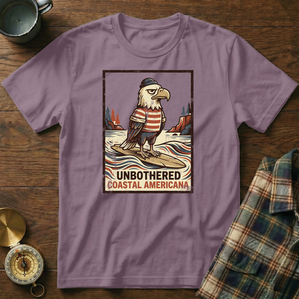 T-shirt with an eagle graphic wearing a hat and striped shirt, surfing. Text reads "UNBOTHERED COASTAL AMERICANA".