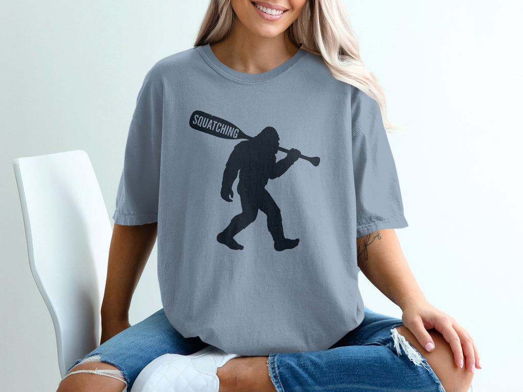 Bigfoot silhouette carrying a paddle labeled "SQUATCHING" on a grey t-shirt.