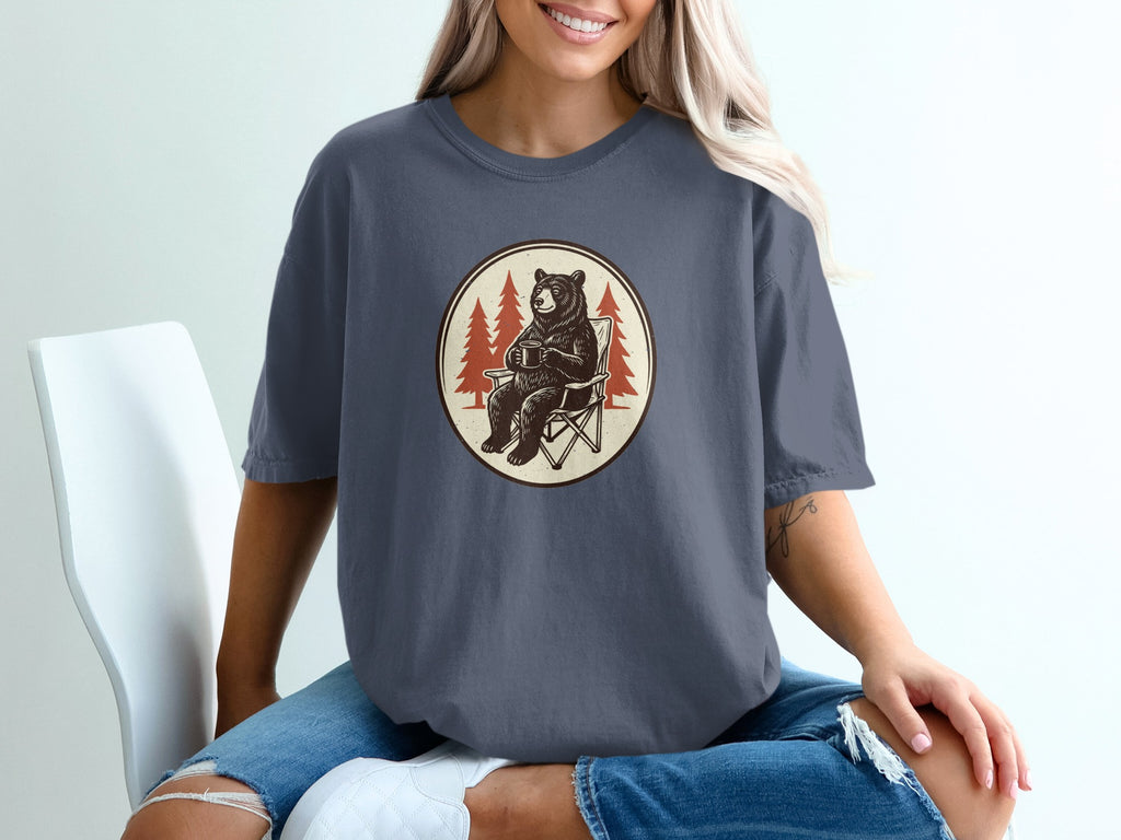 Bear sitting in camp chair holding coffee mug with pine trees in background.