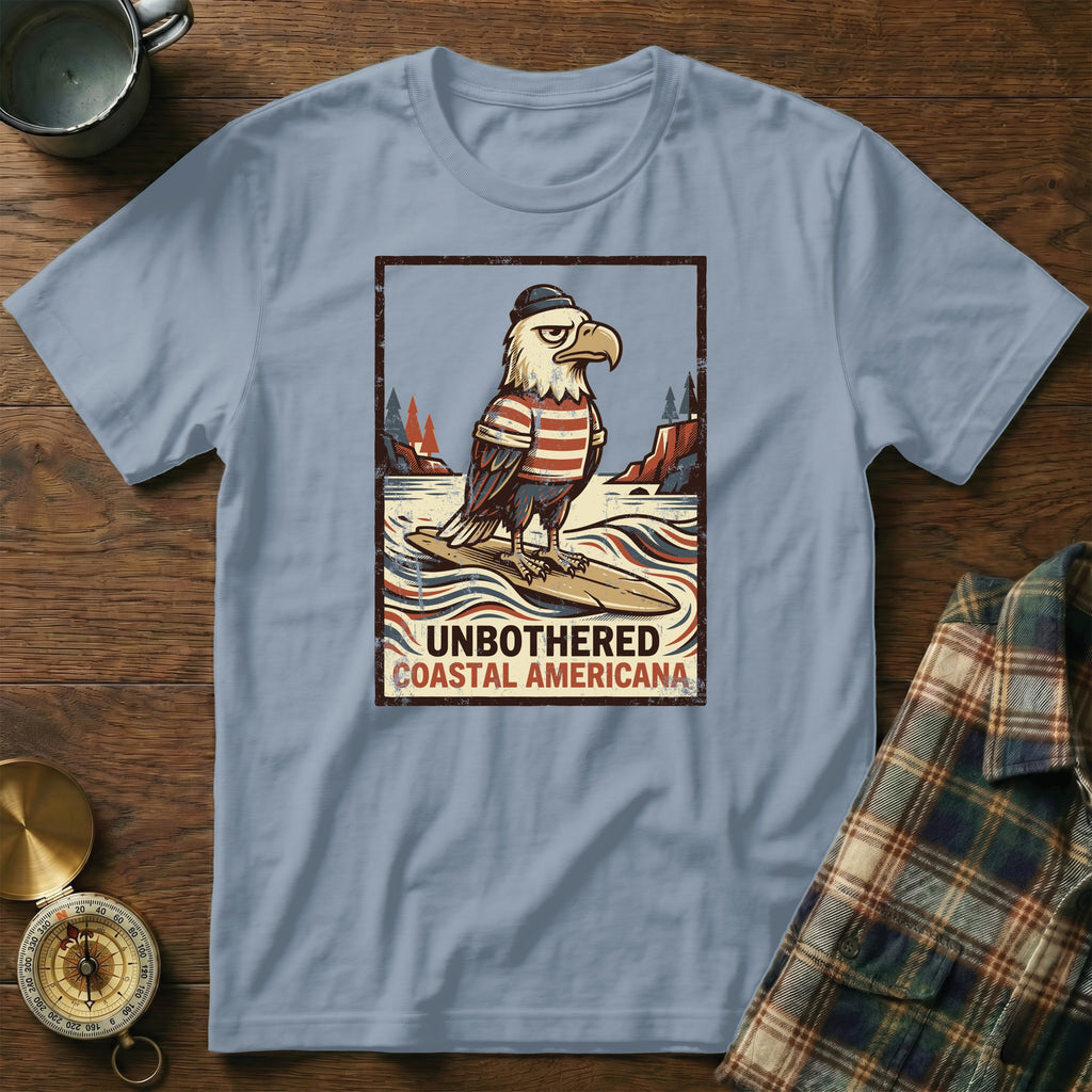T-shirt graphic of an eagle wearing a hat and striped shirt, surfing. Includes text "Unbothered Coastal Americana".