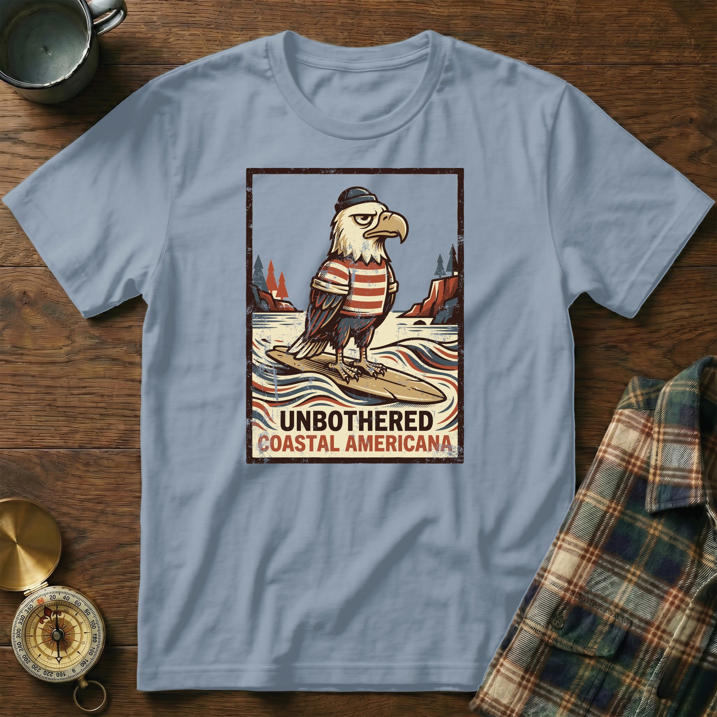 T-shirt graphic of an eagle wearing a hat and striped shirt, surfing. Includes text "Unbothered Coastal Americana".