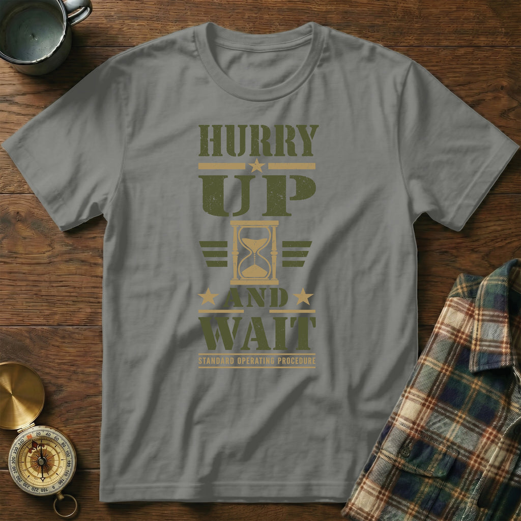 A grey t-shirt printed with "HURRY UP AND WAIT" and an hourglass graphic lies flat on a wooden surface.