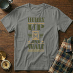 A grey t-shirt printed with "HURRY UP AND WAIT" and an hourglass graphic lies flat on a wooden surface.