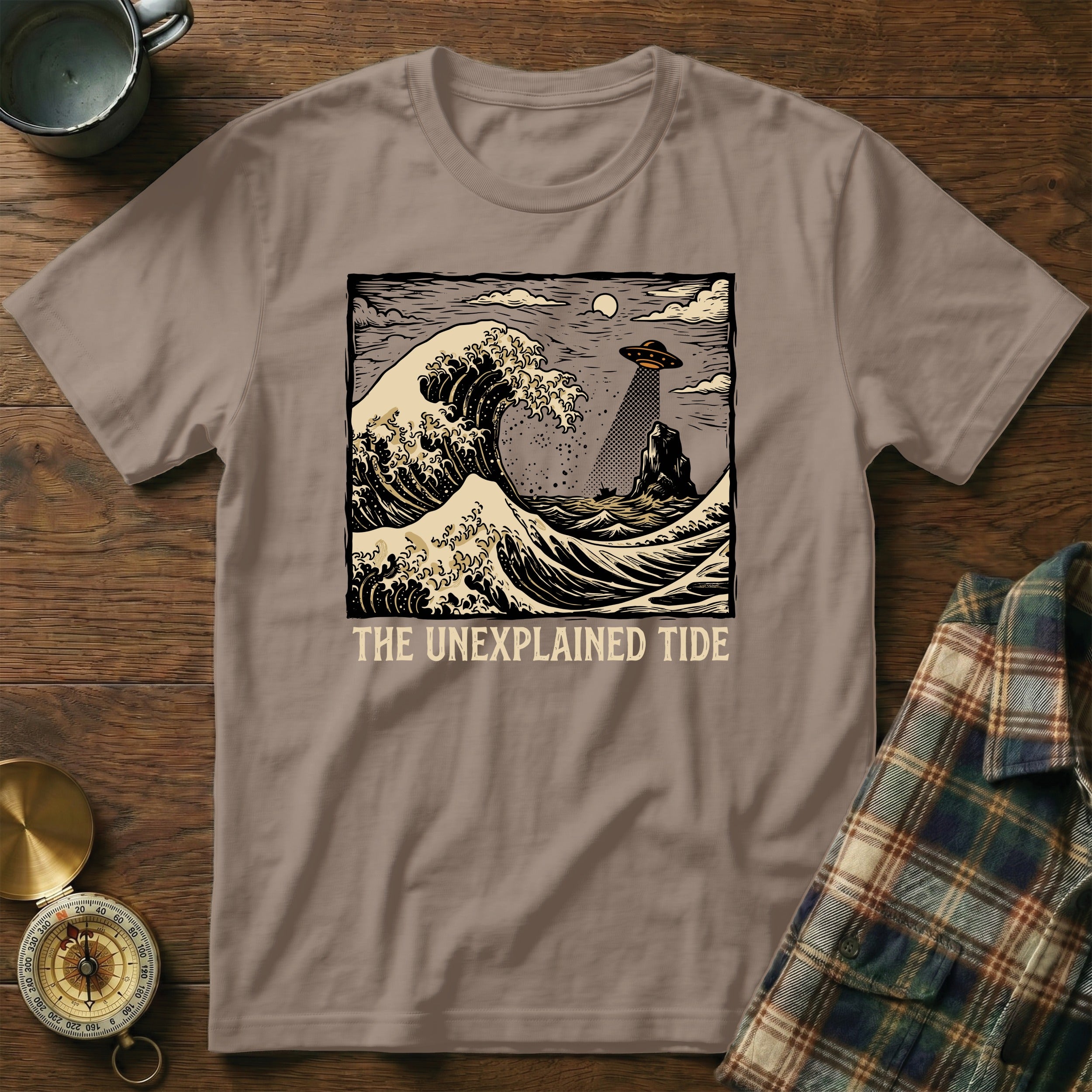 T-shirt graphic: UFO above crashing waves and rocky island, "The Unexplained Tide."