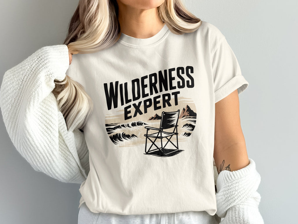 Cream t-shirt with "Wilderness Expert" text and a chair by the sea.