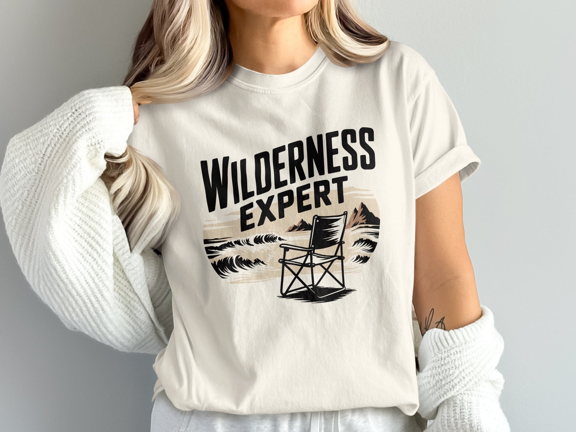 Cream t-shirt with "Wilderness Expert" text and a chair by the sea.