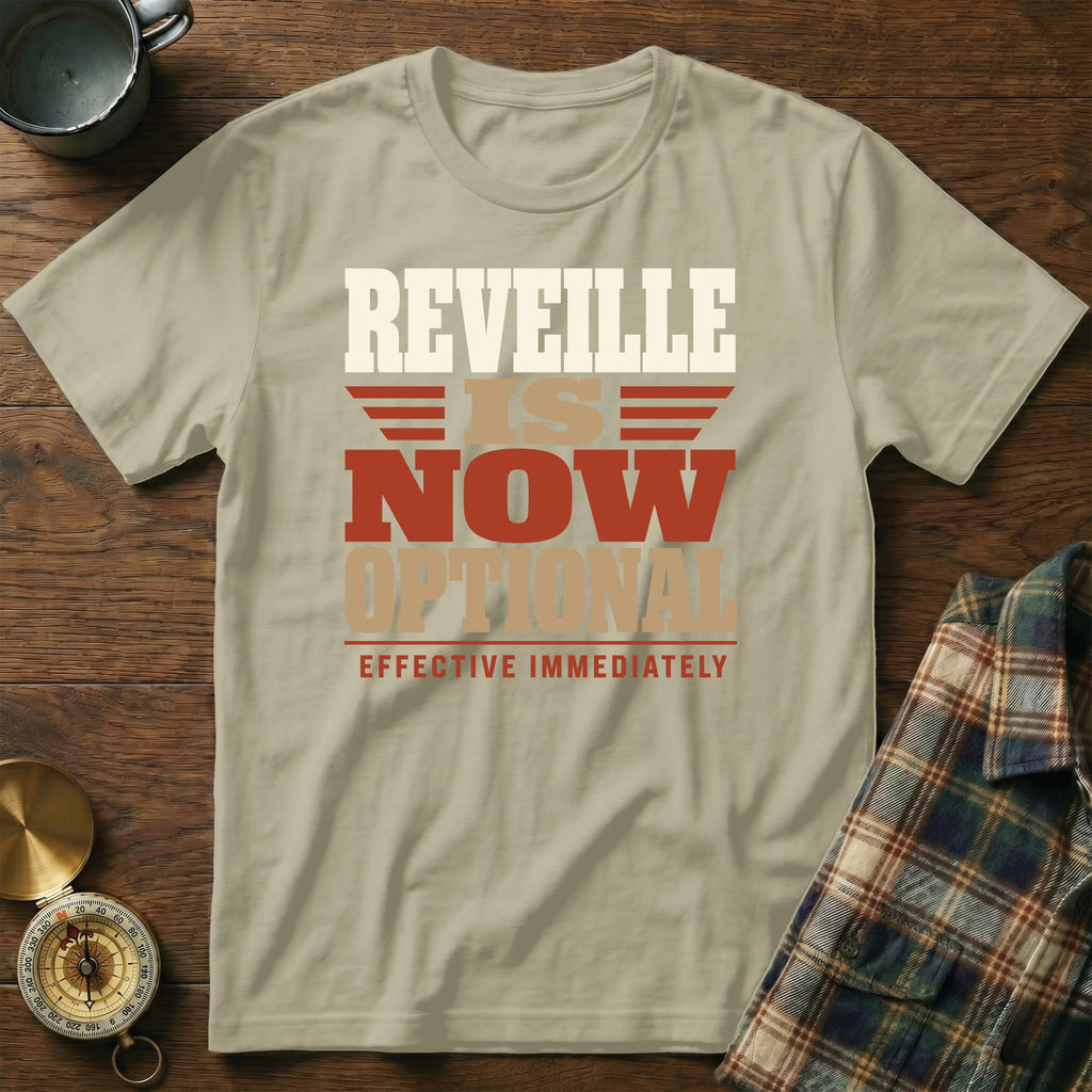 Beige t-shirt featuring the text REVEILLE IS NOW OPTIONAL EFFECTIVE IMMEDIATELY, laid flat on wooden surface.