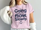 A blonde woman wears a pink crewneck t-shirt featuring the text Going with the flow. Slowly. and an anchor graphic.
