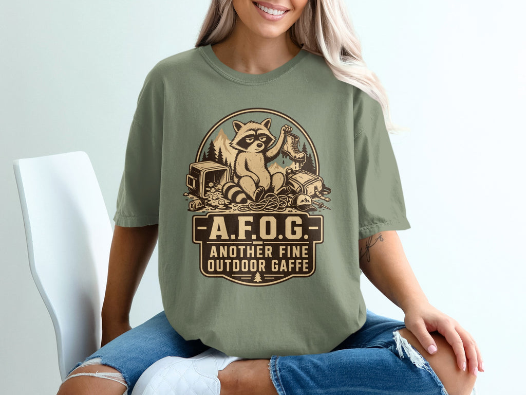 Raccoon holding boot surrounded by camping gear with text "A.F.O.G. ANOTHER FINE OUTDOOR GAFFE".