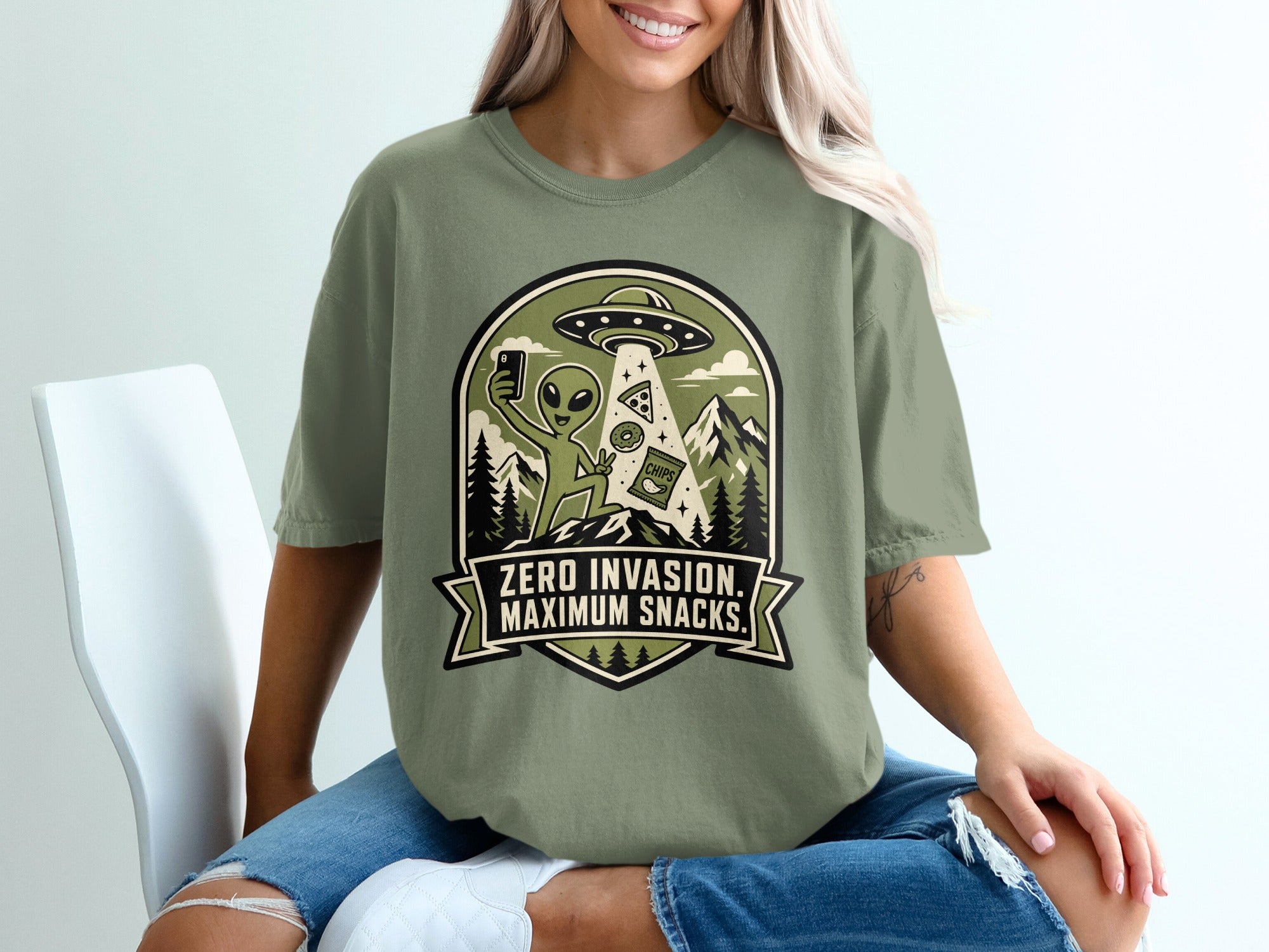 Green t-shirt graphic with alien taking selfie, UFO, snacks, and mountains. Text reads, "ZERO INVASION. MAXIMUM SNACKS."