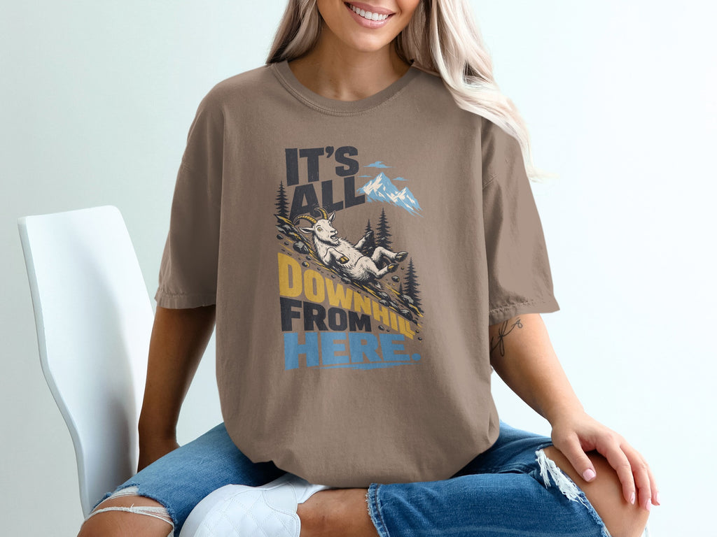 A person wearing a brown t-shirt featuring a mountain graphic and the text It's All Downhill From Here.